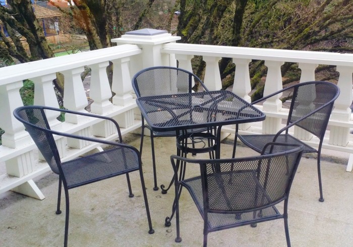 A small outdoor patio with a black metal table and chairs on a concrete floor, surrounded by a white balcony railing and trees in the background.