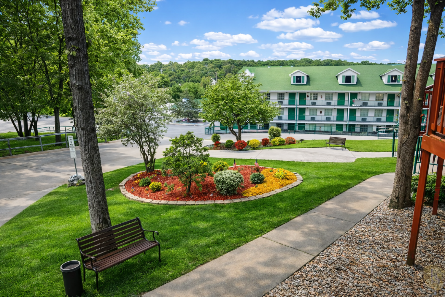 A green-roofed building with white walls and a parking lot surrounded by trees and a garden.