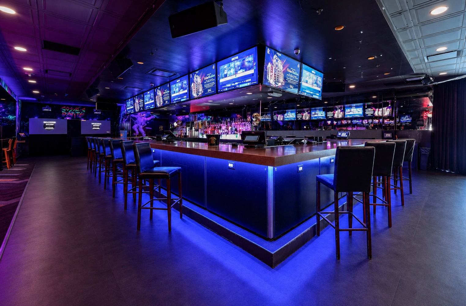 A dimly lit bar with blue lighting, a long counter, and multiple screens.